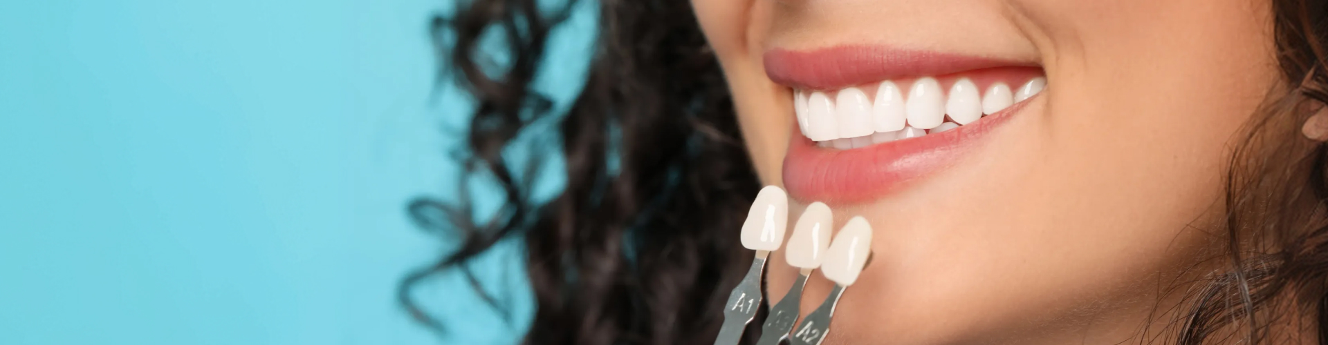 Close-up of a smiling woman holding three tooth shade samples beside her teeth to compare their color.